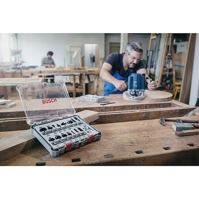 Bosch Fräser-Set Mixed - 15 -tlg., Durchmesser Schaft: 8 mm - Ein Bosch-Fräserset mit verschiedenen Fräsern liegt auf einer Werkbank, während ein Handwerker im Hintergrund eine Oberfräse benutzt.