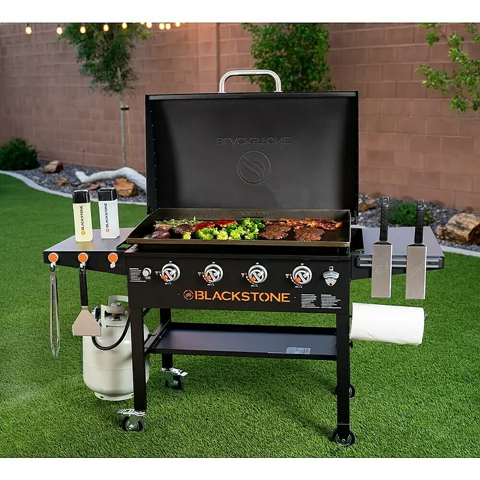 Blackstone Plancha-Grill 36" - Grillfläche (B x T): 91 x 55 cm, 17,5 kW, Anzahl Brenner: 4 Stk. - Ein schwarzer Blackstone-Grill mit Grillgut und Zubehör auf einer Rasenfläche im Freien. Er ist mit praktischen Ablagen und Rollen ausgestattet.