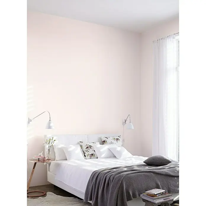 Alpina Wandfarbe Feine Farben - Zarte Romantik, Matt, 2,5 l, Konservierungsmittelfrei - Bett mit weißer Bettwäsche, gemusterten Kissen und einer grauen Strickdecke in einem hellen Schlafzimmer mit rosa Wänden und weißen Lampen.