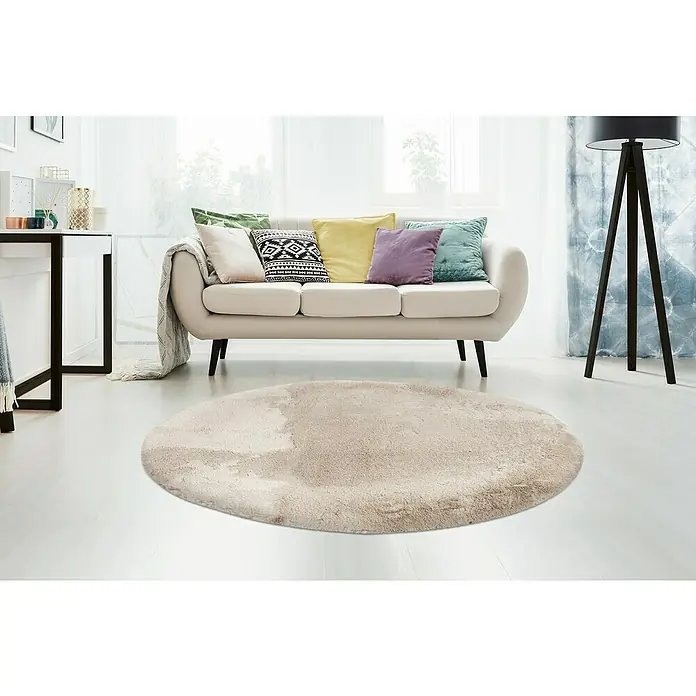 Fellteppich Happy - Light Taupe, Durchmesser: 160 cm, 100 % Polyester (Flor) - Ein cremefarbener runder Teppich liegt auf dem weißen Boden vor einem Sofa mit bunten Kissen. Eine Stehlampe steht rechts.