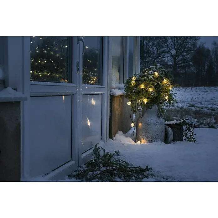 Konstsmide LED-Lichterkette - Außen, Lichtfarbe: Bernstein, Netzbetrieben - Festliche Milchkanne im Schnee: Eine rustikale Milchkanne dient als Vase für Tannenzweige, die mit warmen Lichtern dekoriert sind und winterliche Stimmung verbreiten.
