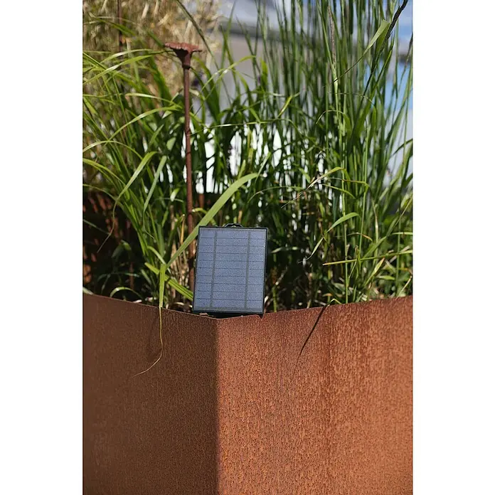 Konstsmide Solar-Batteriebox - 2,5 x 11 x 14 cm - Nahaufnahme eines Solarpanels auf einer rostfarbenen Metallkante vor Gräsern, das nachhaltige Energie und Gartentechnik zeigt.