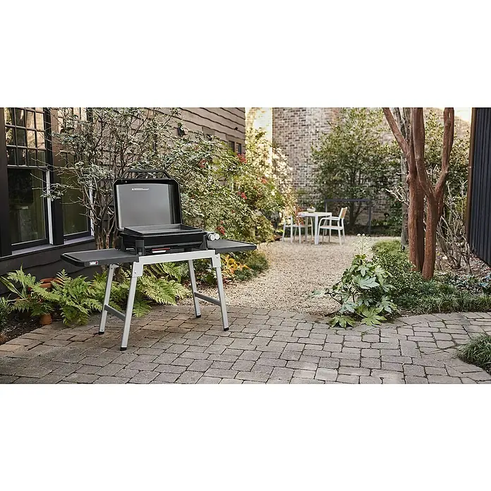 Weber Plancha-Grill Slate GP 56 - Grillfläche (B x T): 56 x 41 cm, 4,7 kW, Anzahl Brenner: 2 Stk. - Nahaufnahme eines Weber Grills mit hochgeklapptem Deckel, der auf einer Terrasse vor einem Garten mit Tisch und Stühlen steht.