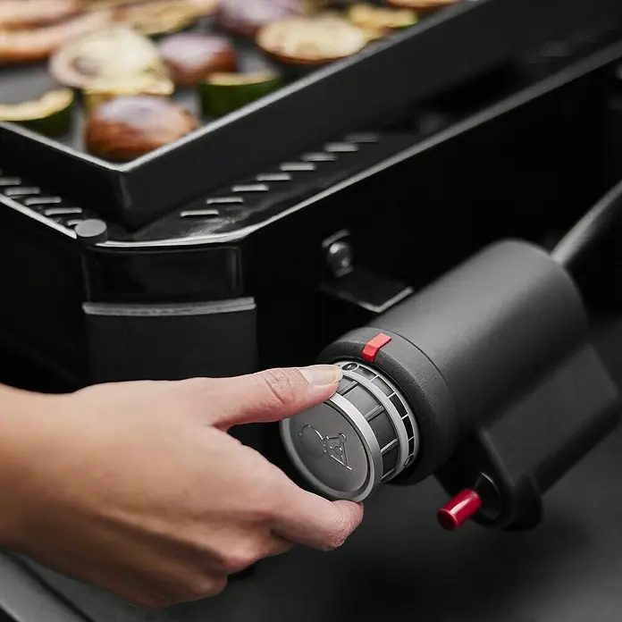 Weber Plancha-Grill Slate GP 43Grillfläche (B x T): 43 x 41 cm, 3,7 kW, Schwarz - Nahaufnahme eines Grills mit Hand am Bedienknopf. Der Grill ist schwarz, während Gemüse auf der Grillfläche zubereitet wird.