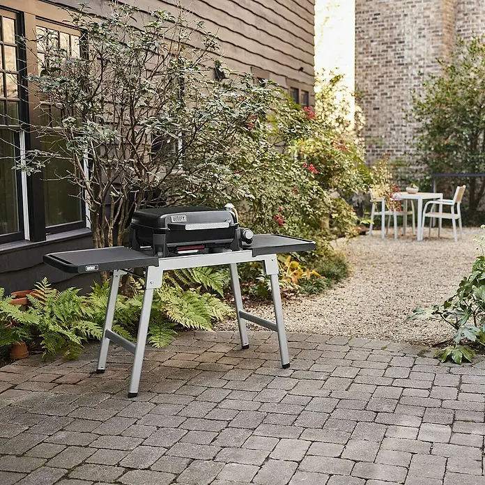 Weber Plancha-Grill Slate GP 43Grillfläche (B x T): 43 x 41 cm, 3,7 kW, Schwarz - Schwarzer Weber Gasgrill auf einem silbernen Standfuß im Außenbereich. Der Grill steht auf einem Steinpflaster vor einem Haus mit Garten.