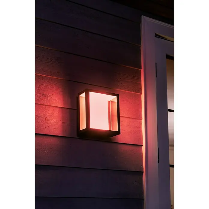 Philips Hue LED-Außenwandleuchte Impress - 8 W, 11,7 x 19 x 24 cm, Schwarz, IP44 - Eine quadratische Außenleuchte in Schwarz strahlt rotes Licht an einer Holzhauswand neben einer Tür aus.