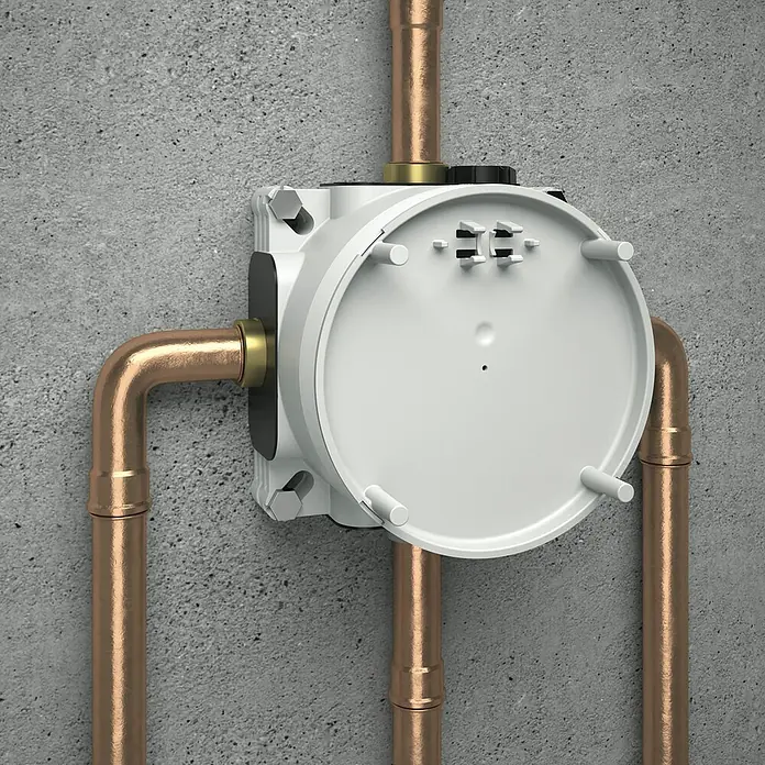 Ideal Standard Unterputzsystem Easy-Box - Einbautiefe: 73 mm - 95 mm - Produktansicht einer Unterputz-Armaturbox mit Kupferrohren an einer Betonwand. Die Box ist weiß und hat einen runden Deckel.