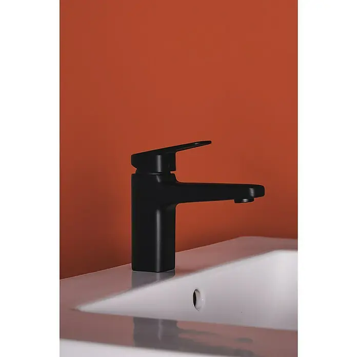 Ideal Standard Ceraplan Waschtischarmatur H90 - Schwarz, Matt, Ohne Ablaufgarnitur - Elegante Nahaufnahme eines mattschwarzen Wasserhahns auf einem weißen Waschbecken, vor einem lebhaften orangefarbenen Hintergrund. Stilvolles Design für moderne Badezimmer.