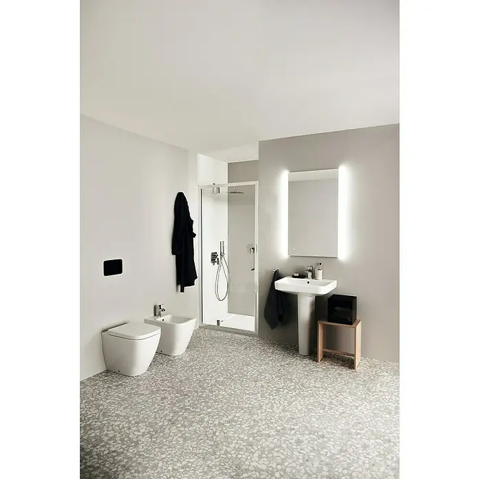 Ideal Standard i.life B Waschtisch60 x 48 cm, Ohne Spezialglasur, Hahnloch: Mitte, Mit Überlauf, Weiß - Ein Badezimmer mit weißen Keramikwaschbecken, Toilette und Bidet, akzentuiert durch einen beleuchteten Spiegel und einem dunklen Bademantel.