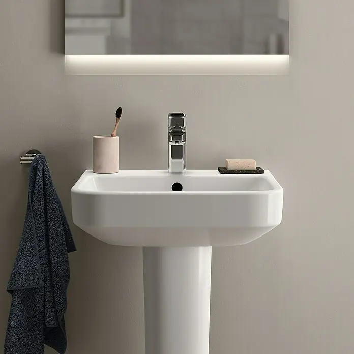 Ideal Standard i.life B Waschtisch - 50 x 44 cm, Ohne Spezialglasur, Hahnloch: Mitte, Mit Überlauf, Weiß - Nahaufnahme eines eleganten weißen Waschbeckens mit Armatur, Zahnputzbecher und Seife, beleuchtet von einem Spiegel.