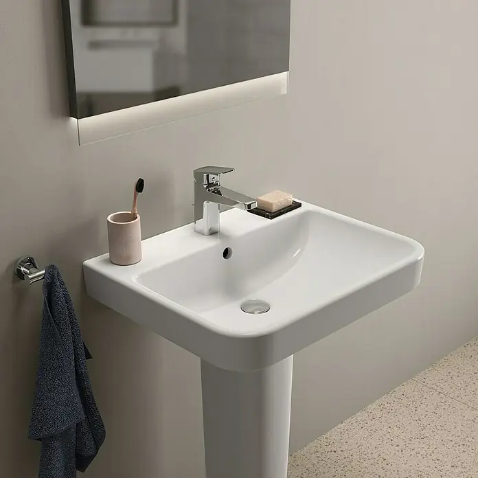 Ideal Standard i.life B Waschtisch - 60 x 48 cm, Ohne Spezialglasur, Hahnloch: Mitte, Mit Überlauf, Weiß - Nahaufnahme eines modernen Waschbeckens mit Armatur, Seife, Zahnbürste und Handtuch vor einer neutralen Wand.