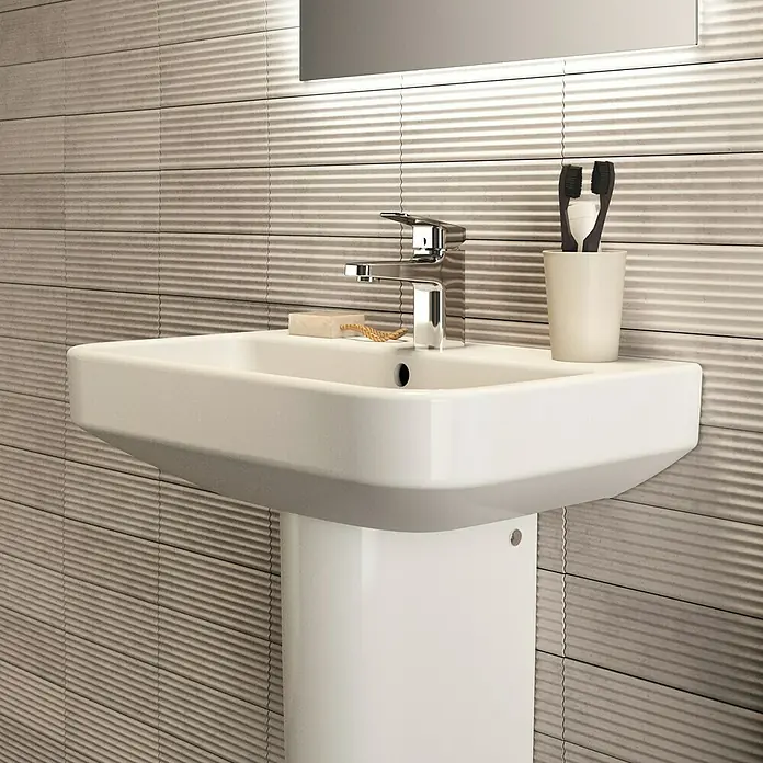 Ideal Standard i.life S Waschtisch - 60 x 38 cm, Ohne Spezialglasur, Hahnloch: Mitte, Mit Überlauf, Weiß - Nahaufnahme eines modernen weißen Waschbeckens mit Armatur, Seife und Zahnbürsten vor einer strukturierten Wand.