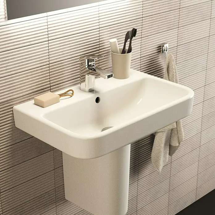 Ideal Standard i.life S Waschtisch - 60 x 38 cm, Ohne Spezialglasur, Hahnloch: Mitte, Mit Überlauf, Weiß - Nahaufnahme eines modernen Waschbeckens mit Armatur, Seife, Zahnbürste und Handtuch in einem Badezimmer mit strukturierten Fliesen.