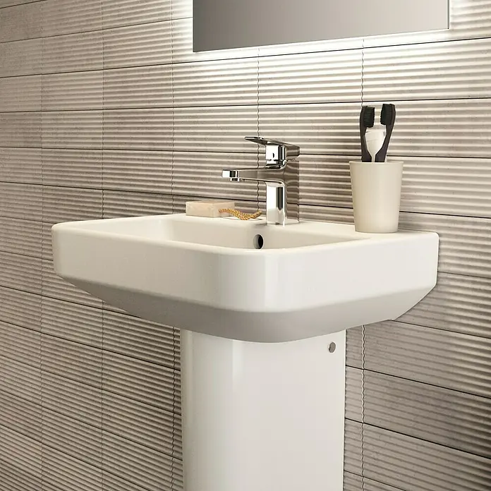 Ideal Standard i.life S Waschtisch - 55 x 38 cm, Ohne Spezialglasur, Hahnloch: Mitte, Mit Überlauf, Weiß - Nahaufnahme eines modernen weißen Waschbeckens mit Armatur, Seife und Zahnbürsten vor einer strukturierten Wand.