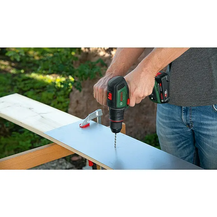Bosch Power for All 18V Akku-Bohrschrauber AdvancedDrill 18V-80 QuickSnap - 18 V, Ohne Akku, 78 Nm, 3 Aufsätze - Ein Mann bohrt mit einem Bosch Akku-Bohrschrauber ein Loch in eine graue Platte, die auf einem Holzbock liegt.