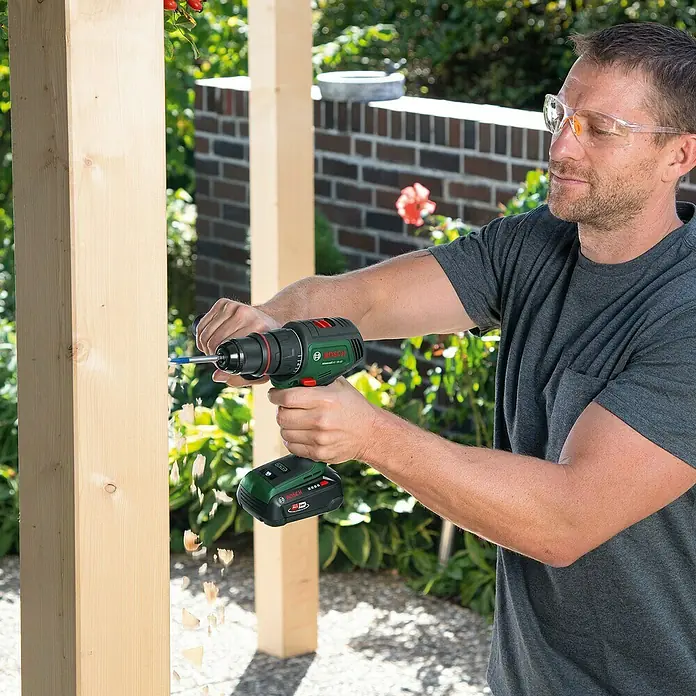 Bosch Power for All 18V Akku-Bohrschrauber AdvancedDrill 18V-80 QuickSnap - 18 V, Ohne Akku, 78 Nm, 3 Aufsätze - Ein Mann bohrt mit einem Akku-Bohrschrauber von Bosch in ein Holzbrett, während er eine Schutzbrille trägt.