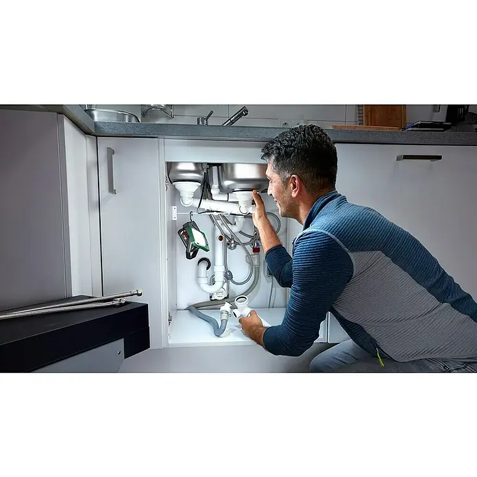 Bosch Power for All 18V Akku-Lampe Easy SpotLight 18V-400 - 18 V, Ohne Akku, 3 Helligkeitsstufen - Ein Handwerker repariert einen Abfluss unter einer Spüle, beleuchtet von einer grünen Arbeitsleuchte, Rohre und Werkzeuge im Blick.