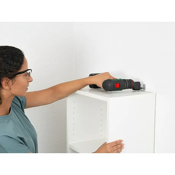 Bosch Power for All 18V Akku-Schlagbohrschrauber Advanced Impact 18V-80 QuickSnap - 18 V, Ohne Akku, 82 Nm, 3 Aufsätze - Eine Frau befestigt mit einem Akkuschrauber einen weißen Schrank an einer weißen Wand. Der Schrank hat mehrere Löcher für Einlegeböden.