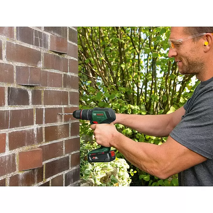 Bosch Power for All 18V Taladro atornillador percutor de batería AdvancedImpact 80 - 18 V, Sin batería, 82 Nm, 3 piezas - Una persona usa un taladro atornillador Bosch verde y negro para perforar una pared de ladrillos marrones al aire libre.