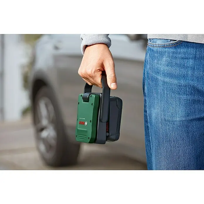 Bosch Power for All 18V Akku-Lampe Easy SpotLight 18V-400 - 18 V, Ohne Akku, 3 Helligkeitsstufen - Nahaufnahme einer Person, die einen grünen Bosch Akku-Adapter mit Griff und Bosch Logo in der Hand hält, im Hintergrund ein Auto.