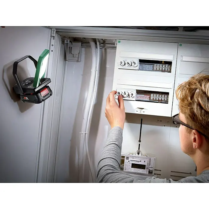 Bosch Power for All 18V Akku-Lampe Easy SpotLight 18V-400 - 18 V, Ohne Akku, 3 Helligkeitsstufen - Eine Akku-Arbeitsleuchte beleuchtet einen Mann, der an einem Sicherungskasten arbeitet, was für gute Sicht bei Elektroarbeiten sorgt.
