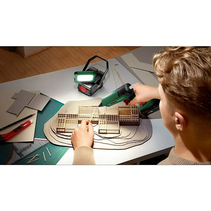Bosch Power for All 18V Akku-Lampe Easy SpotLight 18V-400 - 18 V, Ohne Akku, 3 Helligkeitsstufen - Nahaufnahme eines Mannes, der mit einer grünen Bosch AdvancedGlue 18V Akku-Heißklebepistole ein Architekturmodell klebt. Licht scheint von oben.
