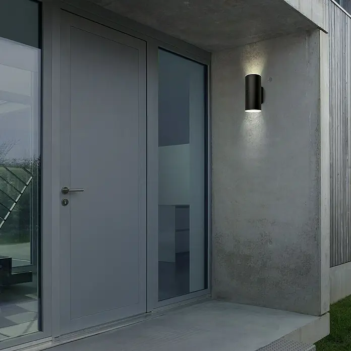 Briloner Akku-Wandleuchte Lima - 15,5 x 7 x 8 cm, Schwarz, Warmweiß, 6 W, IP44 - Eine zylindrische Außenwandleuchte aus dunklem Metall spendet Licht und betont die moderne Architektur des Eingangsbereichs.