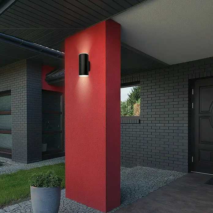 Briloner Akku-Wandleuchte Lima - 15,5 x 7 x 8 cm, Schwarz, Warmweiß, 6 W, IP44 - Eine moderne, schwarze Wandleuchte beleuchtet stilvoll eine rote Säule vor einem Haus. Das Lichtdesign akzentuiert die Architektur.