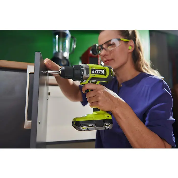 Ryobi ONE+ Akku-Bohrschrauber "Heimwerker-Set" RDD18-2C20TAH - 18 V, 2 Akkus, 2 Ah, 52 Nm - Eine Frau benutzt einen RYOBI Akku-Bohrschrauber, um eine Schraube in ein Möbelstück zu drehen. Schutzbrille und Ohrstöpsel sind getragen.