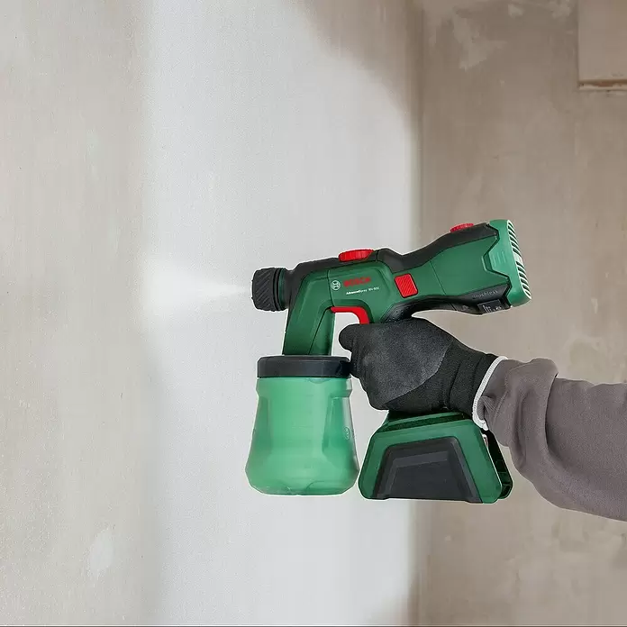 Bosch Power for All 18V Sistema de pulverización de pintura de batería AdvancedSpray 500 - 18 V, 1 batería, 4 Ah, Capacidad de bombeo máx.: 0,5 l/min - Una mano enguantada sostiene una pistola de pintura Bosch verde y negra, rociando pintura blanca sobre una pared.