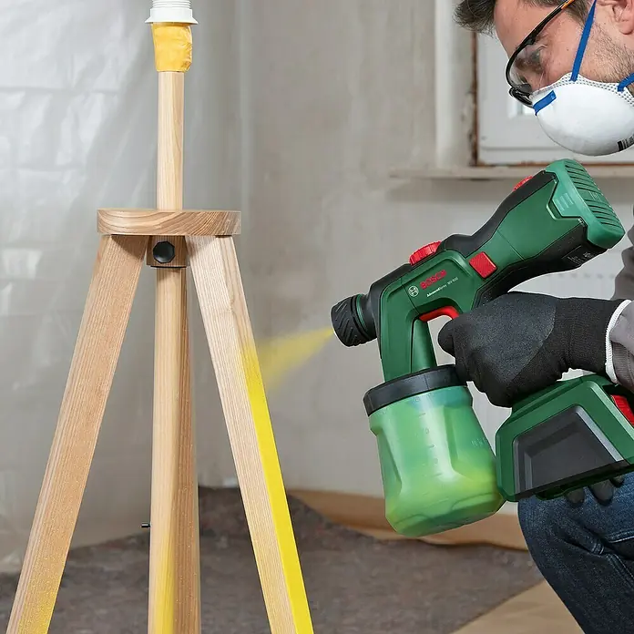 Bosch Power for All 18V Akku-Farbsprühsystem AdvancedSpray 18V-500 - 18 V, Ohne Akku, Max. Fördermenge: 500 l/h - Ein Mann benutzt eine Bosch Farbspritzpistole, um ein hölzernes Stativ zu bemalen. Die Spritzpistole ist grün und schwarz mit einem roten Knopf.