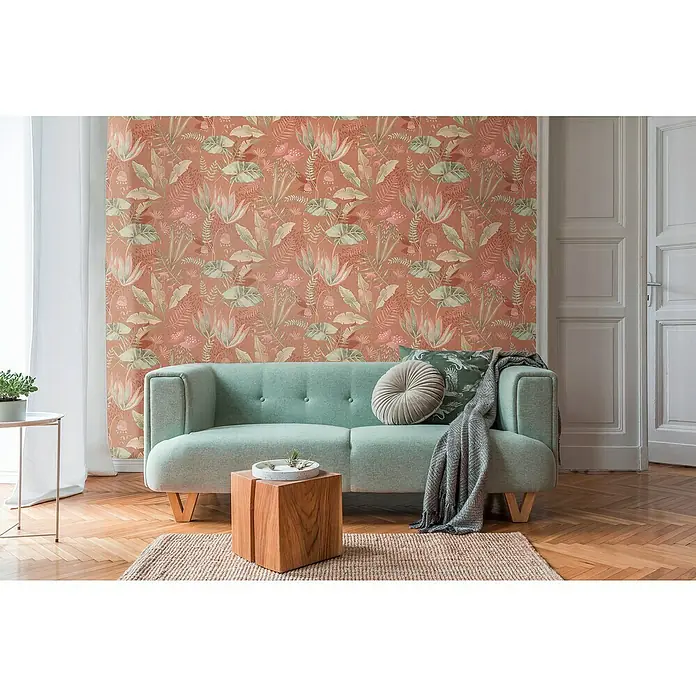 A.S. Création Famous Garden Vliestapete - Rot-Orange-Grün, Floral, 10,05 x 0,53 m - Das Foto zeigt ein grünes Sofa mit Kissen und einer Decke vor einer gemusterten Wand. Die Szene wird durch einen Holzhocker und Teppich ergänzt.