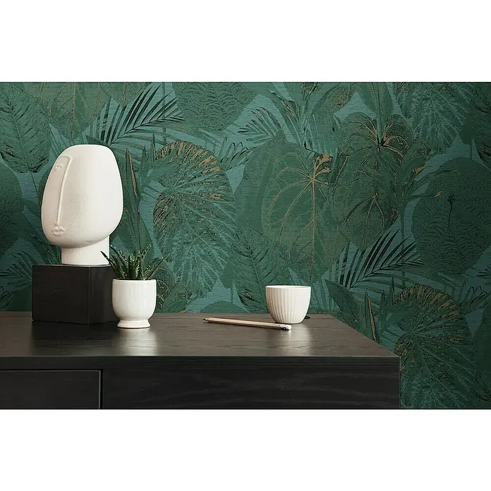 A.S. Création Famous Garden Vliestapete Dschungel - Petrol-Grün-Gold, Floral, 10,05 x 0,53 m - Nahaufnahme eines stilvollen Interieurs mit einer dekorativen Gesichtsskulptur und einem kleinen Kaktus vor einem tropischen Blattmuster.