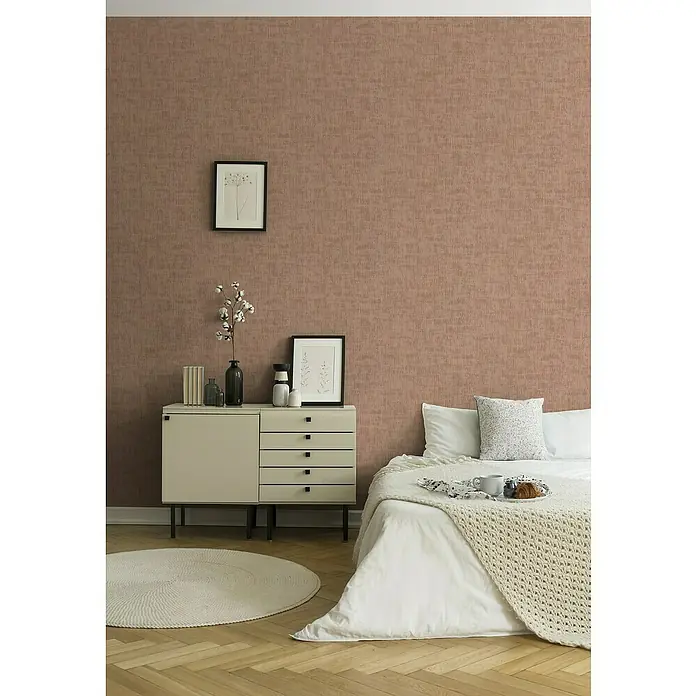 A.S. Création Jade 2 Vliestapete - Rosa, Uni, 10,05 x 0,53 m - Das Schlafzimmer verfügt über eine Kommode und ein Bett mit Kissen und Decke vor einer strukturierten Wand. Eine runde Matte liegt auf dem Boden.