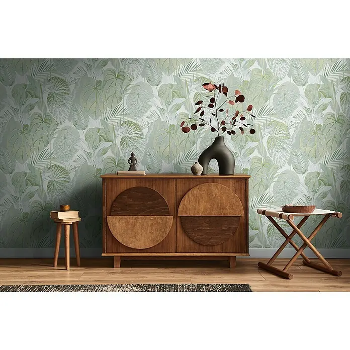 A.S. Création Famous Garden Vliestapete Dschungel - Grün-Weiß-Grau, Floral, 10,05 x 0,53 m - Elegante Anrichte aus Holz mit runden Türen, verziert mit einer dunklen Vase und einer kleinen Statue, vor einer botanischen Tapete.