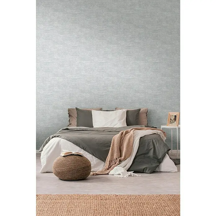 A.S. Création Stories of Life Vliestapete - Grau-Silber, Uni, 10,05 x 0,53 m - Ein Schlafzimmer mit einem Bett, mehreren Kissen, einer Decke und einem Hocker. Der Teppich ist braun und die Wand ist hellgrau.