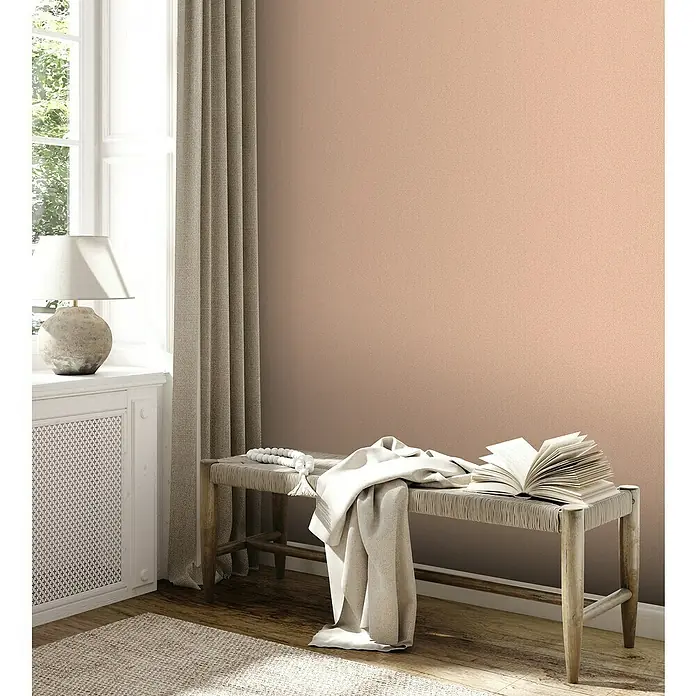 A.S. Création Jade 2 Vliestapete - Rosa-Orange, Uni, 10,05 x 0,53 m - Einladender Raum mit einer hellrosa Wand, einem bequemen Holzbänkchen, auf dem ein Buch und eine Decke liegen.