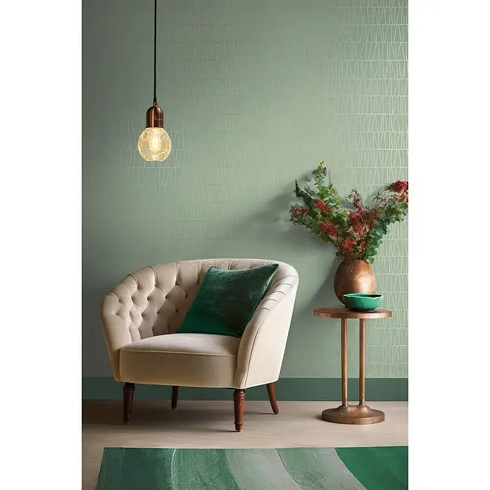 A.S. Création Jade 2 Vliestapete - Goldgrün, Grafisch, 10,05 x 0,53 m - Eine elegante Wohnzimmerinszenierung mit einem beigen Ohrensessel, einem grünen Kissen und einer stilvollen Stehleuchte.