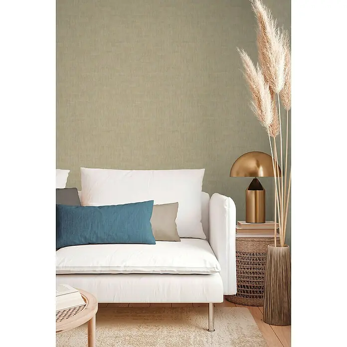A.S. Création Jade 2 Vliestapete - Beige-Taupe, Uni, 10,05 x 0,53 m - Wohnzimmer-Arrangement mit einem eleganten weißen Sofa, akzentuiert durch blaue und graue Kissen, neben einer stilvollen goldenen Lampe und dekorativem Pampasgras.