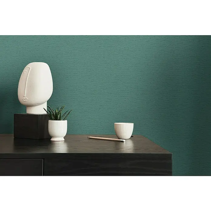 A.S. Création Famous Garden Vliestapete - Petrol-Grün, Uni, 10,05 x 0,53 m - Elegante Nahaufnahme einer Kommode mit Deko-Objekten, darunter eine Skulptur und eine kleine Pflanze in einem weißen Topf, vor einer grünen Wand.