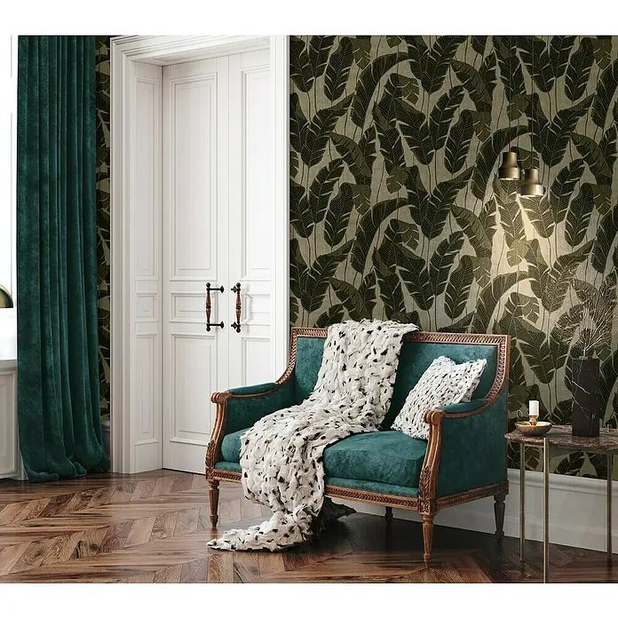 A.S. Création Jade 2 Vliestapete Blätter - Schwarzbeige, Floral, 10,05 x 0,53 m - Elegantes Interieur mit einem smaragdgrünen Sofa, das mit einer flauschigen Decke und einem Kissen dekoriert ist, vor einer kunstvollen Tapete.