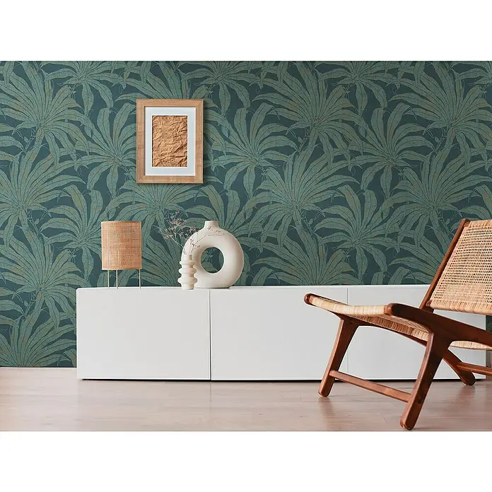 A.S. Création Jade 2 Vliestapete Pflanzen - Petrol-Gold-Blau, Floral, 10,05 x 0,53 m - Wohnzimmer mit einem weißen Sideboard, einer Lampe, einer Vase, einem Bild und einem hölzernen Loungesessel vor einer dunkel gemusterten Dschungeltapete.