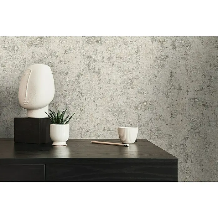 A.S. Création Jade 2 Vliestapete - Weiß-Taupe-Rosa, Uni, 10,05 x 0,53 m - Nahaufnahme eines Dekors mit einer abstrakten Skulptur, einer kleinen Pflanze und einem Bleistift auf einer schwarzen Kommode vor einer strukturierten Wand.