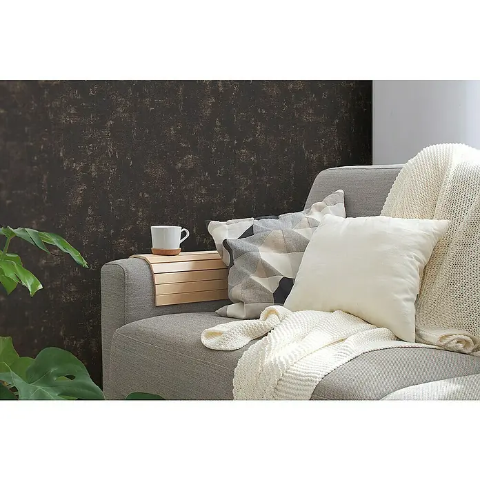 A.S. Création Jade 2 Vliestapete - Schwarz-Gold, Uni, 10,05 x 0,53 m - Gemütliches Wohnzimmer mit grauem Sofa, Zierkissen und Strickdecke. Eine Tasse steht auf einem Armlehnbrett. Zimmerpflanze im Vordergrund.