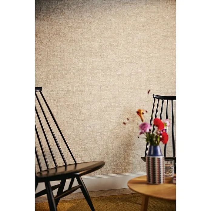 A.S. Création Stories of Life Vliestapete - Beige-Weiß-Gold, Uni, 10,05 x 0,53 m - Nahaufnahme einer strukturierten, beigen Wand mit einem dunklen Holzstuhl und einem runden Tisch mit Blumenvase.