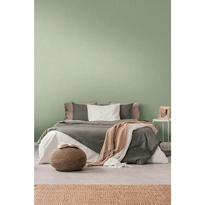A.S. Création Jade 2 Vliestapete - Goldgrün, Grafisch, 10,05 x 0,53 m - Schlafzimmer mit Bett, Pouf und Teppich. Das Bett ist mit grauer und weißer Bettwäsche bezogen, ein beiger Überwurf liegt darüber.