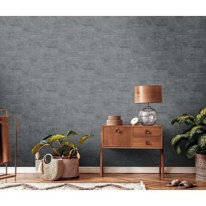 A.S. Création Stories of Life Vliestapete - Schwarz-Grau-Gold, Uni, 10,05 x 0,53 m - Elegantes Interieur mit einer grauen Wandtapete, die dem Raum eine stilvolle Textur verleiht. Ein Holztisch und Pflanzen ergänzen das Ambiente.