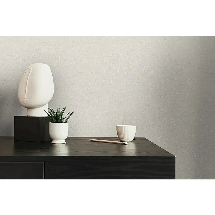 A.S. Création Jade 2 Vliestapete - Creme-Beige, Uni, 10,05 x 0,53 m - Elegante Nahaufnahme einer schwarzen Kommode mit einer stilvollen Gesichtsskulptur, einem Kaktus, einer Tasse und einem Bleistift.