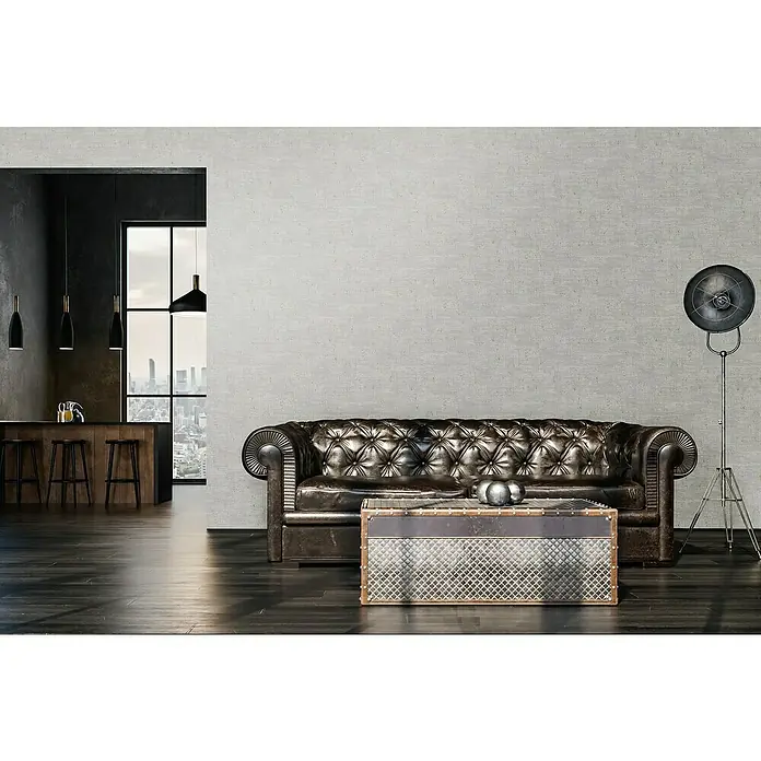 A.S. Création Stories of Life Vliestapete - Grau-Creme-Gold, Uni, 10,05 x 0,53 m - Ein Chesterfield-Ledersofa mit einem Metall-Couchtisch im Vintage-Stil vor einer grauen Wand und einer Stehlampe.