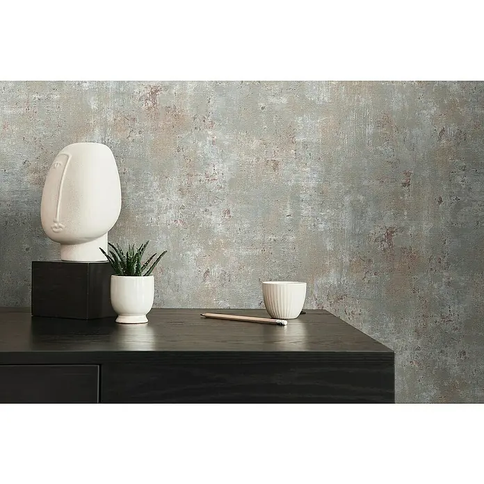 A.S. Création Stories of Life Vliestapete Patina - Grau-Blau-Bronze, Betonoptik, 10,05 x 0,53 m - Dekorative Nahaufnahme eines Tisches mit Keramikkopf, Topfpflanze, Tasse und Bleistift vor gemusterter Wand, die ein minimalistisches Ambiente erzeugt.